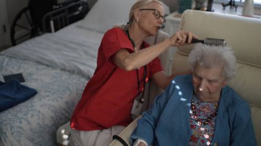 Bakıcı, yaşlı bir kadına el aynasında yansımasını incelerken saçı için nazikçe yardım eder. Sıcak, davetkar atmosfer onların etkileşimini güçlendirir..