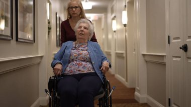 Bir bakıcı yaşlı bir kadına yardım ediyor. Tekerlekli sandalyedeki yaşlı bir kadına. Gündüz vakti, iyi aydınlatılmış bir huzur evinin koridorunda..