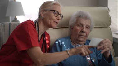 Bakıcı yaşlı bir kadınla iletişim kurar, hatıraları canlandırmak için bir fotoğrafa işaret eder. Bağlantılar, sağlık merkezlerindeki kişisel bakım merkezlerini vurguluyor.