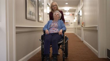 Bir bakıcı, tekerlekli sandalyedeki yaşlı bir kadına bir sağlık merkezinin aydınlık koridorunda yardım eder. Etkileşim sırasında bağlanma ve destek sağlar..