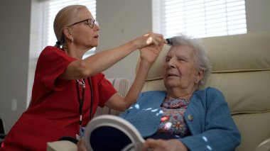 Bakıcı, yaşlı bir kadının saçlarını nazikçe şekillendirir. O da rahat bir şekilde sandalyede oturur. Güneş ışığı pencereden süzülerek sıcak bir atmosfer yaratır..