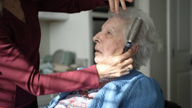 Bakıcı, yaşlı bir kadının saçlarını parlak ve rahat bir odada tarar. Kadın bir sandalyede dinleniyor, öğleden sonra şımartma anının tadını çıkarıyor..