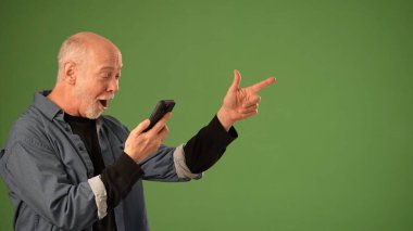 Olgun bir adam akıllı telefonuyla neşeli bir şekilde etkileşime girer ve sağlam yeşil bir zemin üzerinde coşkuyla işaret eder. Onun ifadesi şaşkınlık ve mutluluk taşıyor..