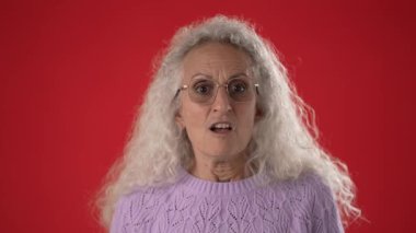 50 'li ya da 60' lı yaşlarda beyaz saçlı ve gözlüklü olgun bir kadın canlı kırmızı bir arka planla çevrili iken şaşkınlık ya da heyecanı tutkuyla ifade eder..