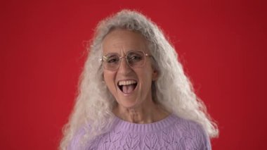Orta yaşlı, gözlüklü ve beyaz saçlı bir kadın mutlu bir şekilde dalgalanıyor. Sıcak gülüşünü, neşeli davranışlarını artıran canlı kırmızı bir arka planda sergiliyor..