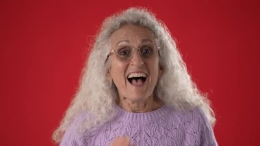 50 ya da 60 yaşlarında olgun bir kadın gözlük takarken sevinçle bağırıyor, beyaz kıvırcık saçlarını ve canlı kişiliğini kırmızı bir arka planda sergiliyor..