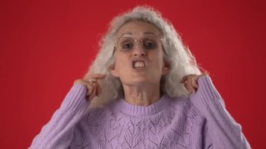 Çekici özellikleri ve gözlükleri olan orta yaşlı bir kadın gülümseyerek kulaklarını şakayla kapatıyor. Canlı kırmızı bir arkaplanda, canlı bir atmosfer yaratıyor..