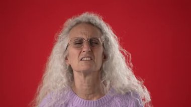 Beyaz saçlı ve gözlüklü orta yaşlı bir kadın ellerini kırmızı bir ortamda kaldırıp duygularını yansıtan neden ben olduğumu soruyor..