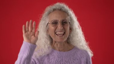 Beyaz kıvırcık saçlı, gözlüklü, gülümseyen ve el sallayan olgun bir kadın. Canlı kırmızı arka plan, bu canlı portredeki neşeli ifadesine enerji katıyor..