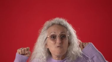 Beyaz saçlı ve gözlüklü orta yaşlı bir kadın kollarını kaldırıp gülümseyerek başarı ve heyecan ifade ediyor. Açık mor bir kazak giyiyor ve anın tadını çıkarıyor..