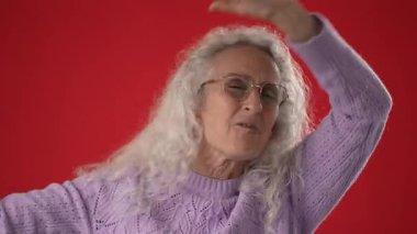 Çekici özellikleri ve gözlükleriyle orta yaşlı bir kadın kollarını kaldırıyor, özgüvenini ve başarısını cesur kırmızı arka planda sergiliyor..