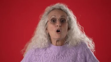 Beyaz saçlı, gözlüklü, orta yaşlı bir kadın sürpriz ve öfkeyle ellerini kaldırıyor. Mor bir kazak giyiyor. Parlak kırmızı arka plan onun anlamlı özelliklerini arttırıyor..