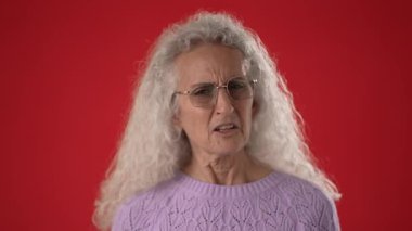 50 ya da 60 yaşlarında orta yaşlı bir kadın der ki Kim ben onun doğal güzelliği beyaz kıvırcık saçlı ve şık gözlüklü, lavantalı süveter giyen canlı kırmızı bir arka planda.