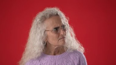 Orta yaşlı, beyaz saçlı ve gözlüklü bir kadın parmağını dudaklarına dayayıp sessizlik işareti yapıyor..