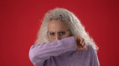 Gözlük takan orta yaşlı bir kadın ve soğuk algınlığından muzdarip mor bir kazak. Grip, canlı kırmızı bir arkaplan karşısında duruyor. Duygularını iletiyor..