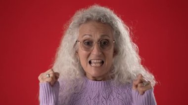 Beyaz saçlı ve gözlüklü orta yaşlı bir kadın üzgün, mutsuz, yumruklarını sıkan ve canlı kırmızı bir zeminde anlamlı bir yüz ifadesi sergiliyor..