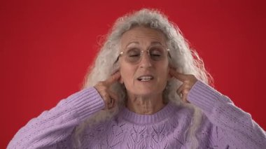 Beyaz saçlı ve gözlüklü orta yaşlı bir kadın açık mor bir kazak giyerken kulaklarını kapatarak şakacı bir poz veriyor. Kalın kırmızı arkaplan enerji katar.