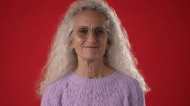 50-60 yaşlarında, gözlüklü, kıvırcık beyaz saçlı açık mor bir kazak giyerken gülümseyen orta yaşlı bir kadın. Arkaplan parlak kırmızı.