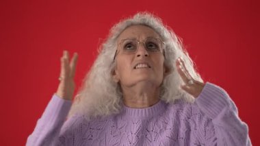 Gözlük takan orta yaşlı bir kadın ve açık mor bir süveter görünür bir şekilde kızgın, hayal kırıklığına uğramış, iki eliyle canlı kırmızı bir arkaplana karşı başını tutuyor..