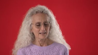 Orta yaşlı bir kadın güzel anıların hayalini kuruyor gözlük takıyor ve mor bir süveter giyiyor sıcak bir şekilde gülümsüyor. Beyaz saçları zarifçe dökülüyor, canlı kırmızı bir arkaplan karşısında çekici bir portre oluşturuyor..