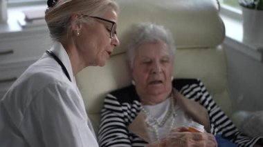 Olgun bir kadın rahat bir koltukta otururken bir bakıcı ilaçlarını açıklıyor. Sıcak bir yuva ortamında ortam sakin ve destekleyici..