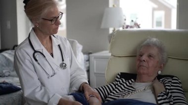 Olgun bir kadın, ev ziyareti sırasında bir sağlık görevlisiyle el ele tutuşarak sandalyesinde rahatlar. Atmosfer sakin ve şefkatli..