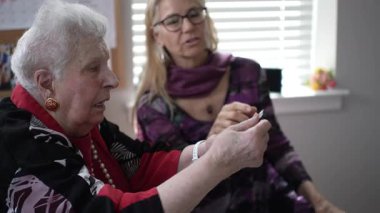 Yaşlı bir kadın kızıyla otururken uzaktan kumanda kullanmaya odaklanır, sıcak aydınlatılmış bir oturma odasında sıcak bir ziyaretin tadını çıkarır..