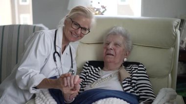 Bakıcı ev ziyareti sırasında yaşlı bir kadınla iletişim kurar, rahat bir oturma odasında rahatlık ve arkadaşlık teklif eder..