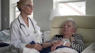 Olgun bir kadın rahat bir sandalyede dinlenirken bir sağlık uzmanı ev ziyareti sırasında sağlığını dikkatle kontrol ediyor ve rahatlatıcı bir atmosferi teşvik ediyor..