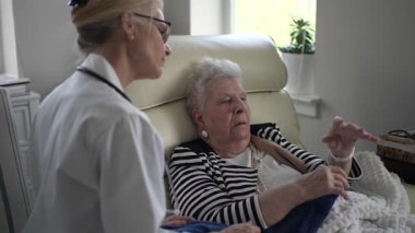 Yaşlı bir kadın evde bakıcısıyla sohbet eder, bir sandalyede rahat bir şekilde otururken düşüncelerini paylaşır ve rahat atmosferin tadını çıkarır..