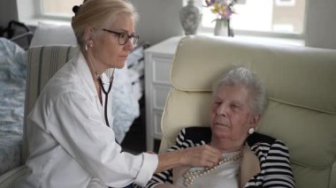 Olgun bir kadın rahat evindeki bir hemşireden sağlık kontrolü alır. Hemşire steteskop kullanırken kadın rahat bir şekilde koltukta oturuyor..