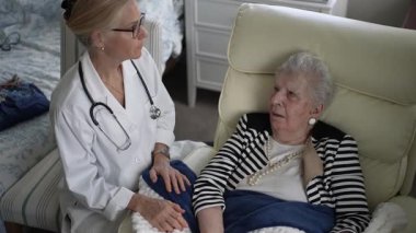 Yetişkin bir kadın evdeki bir sandalyede rahatça otururken, bir sağlık uzmanı onunla konuşarak ziyaret sırasında arkadaşlık ve tıbbi tavsiyelerde bulunuyor..