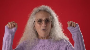 Gülümseyen orta yaşlı, beyaz saçlı ve gözlüklü bir kadın ellerini havaya kaldırarak mutluluğunu ifade eder..