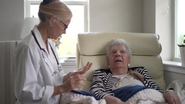 Bakıcı, evinde rahat bir koltukta oturan yaşlı bir kadınla dikkatle konuşur. Ziyaret sırasında rahatlık ve destek sağlar..