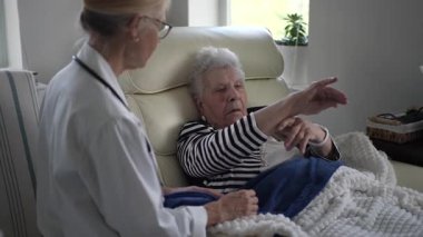 Olgun bir kadın evindeki koltuğunda rahatça oturur ve dikkatle dinleyen bir bakıcı ile konuşur. Bir anlık bağ ve bakım paylaşıyorlar..