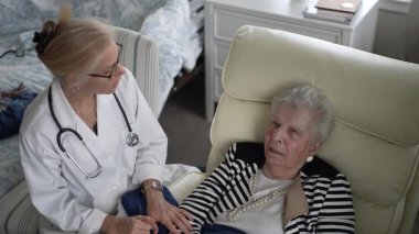 Bir sağlık hizmetleri uzmanı, yaşlı bir kadına evinde bakım sağlıyor. Oturma odasında rahatça otururken sohbet ediyorlar..