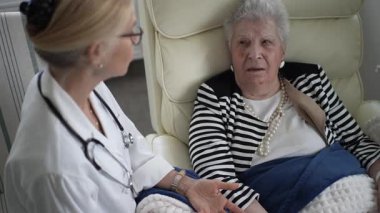 Olgun bir kadın, bir sağlık uzmanı sağlığını tartışırken rahat bir şekilde bir sandalyeye oturur. Konforlu ev atmosferi, kişisel bakımın önemini vurguluyor.