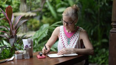 Orta yaşlı bir kadın yemyeşil bir kafeteryada oturmuş tatilin tadını çıkarırken defterini hazırlıyor. Dijital göçebe yaşam tarzını somutlaştırıyor..