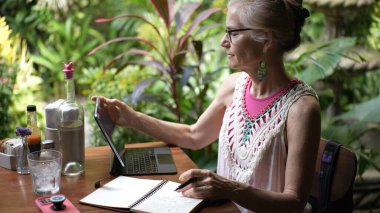 Yeşillik bir kafede, gözlüklü olgun bir kadın, notları yazarken tabletle meşgul olur, tatildeki dijital göçebenin ruhunu somutlaştırır..