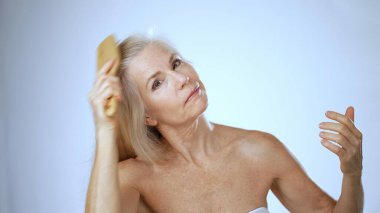 Olgun bir kadın, uzun, dalgalı saçlarını tararken gülümseyen sakin bir spa ortamında oturuyor. Huzurlu atmosfer onun mutluluğunu arttırıyor..