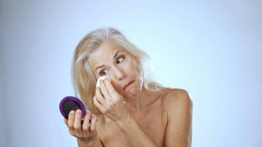 Olgun bir kadın spa gününün tadını çıkarırken mutlu bir şekilde cilt bakımı yapıyor. Gülümsüyor ve aynadaki yansımasına odaklanıyor. Basit beyaz bir arka plana karşı..