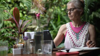 Olgun bir kadın bir kafede oturup tabletine odaklanıyor. Enerjik yeşillik, dinlendirici bir ortamda iş ve tatil dengesini sağlarken huzurlu bir atmosfer yaratıyor..