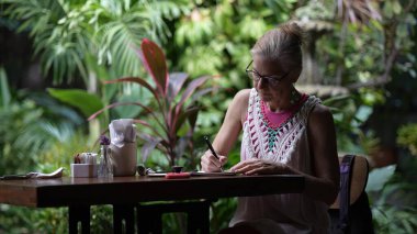 Orta yaşlı bir kadın kafeterya masasında oturuyor, akıllı telefonuna odaklanmış, yemyeşil şeylerle çevrili. Huzurlu bir ortamda iş ve boş zamanı dengeliyor..
