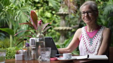 Orta yaşlı bir kadın yemyeşil bir kafenin etrafında çalışırken gülümsüyor. Dijital göçebe yaşam tarzına dalmışken kahve içmekten zevk alıyor..