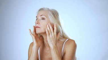 Uzun saçlı olgun bir kadın gülümserken nemlendirici sürüyor. Parlak, sakin bir spa ortamında rahat ve mutlu görünüyor..