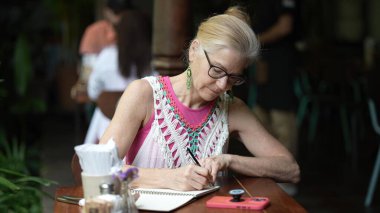 Orta yaşlı bir kadın bir kafe masasında oturuyor, kendi yazısıyla meşgul. Çevresi yeşilliklerle çevriliyken, dijital göçebe olarak tatildeyken işinden zevk alıyor..