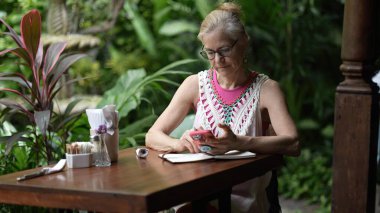 Yetişkin bir kadın kafeterya masasında oturuyor, telefonuna ve defterine odaklanmış, yemyeşil şeylerle çevrili. Tatili boyunca iş ve dinlenme dengesi kurar..