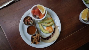 Orta yaşlı bir kadın, kafeterya masasında oturmuş, tatil sırasında renkli bir tabak kahvaltılık yemeğin tadını çıkarıyor..