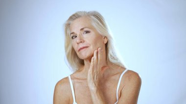 Kaplıcada olgun bir kadın sıcak bir gülümsemeyle mutluluğu ifade eder. Nazikçe yanağına dokunuyor, parlak derisini açık bir arka planda gösteriyor..