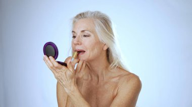 Mutlu, olgun bir kadın sakin bir spa atmosferiyle çevrili aynaya bakarken ruj sürer. Neşeli ifadesi rahatlama ve kendine güveni yansıtıyor..